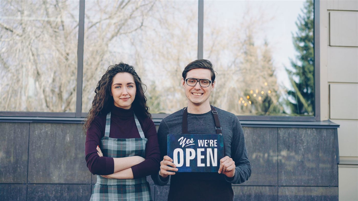 Two small business owners holding a 'Yes We're Open' sign outside their shop